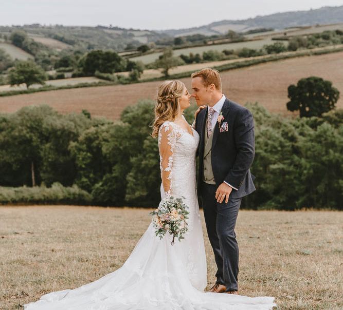 Bride in Lace Sincerity Bridal Wedding Dress and Groom in Blue Ted Baker Suit Embracing in Field