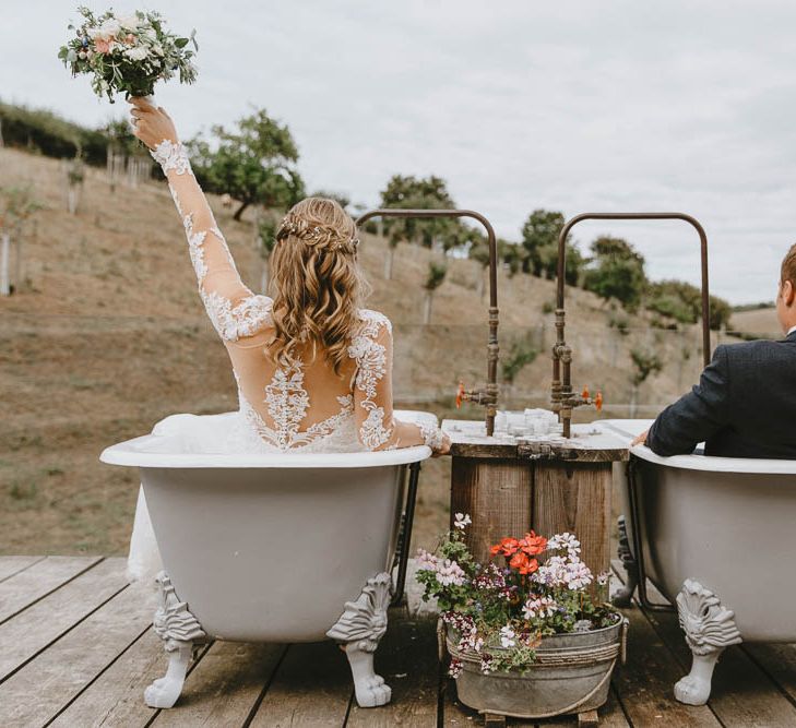 Bride in Lace Sincerity Bridal Wedding Dress and Groom in Blue Ted Baker Suit Sitting in Twin Rolltop Baths on The Old Piggyery Veranda at Devon Wedding Venue Windout Barn