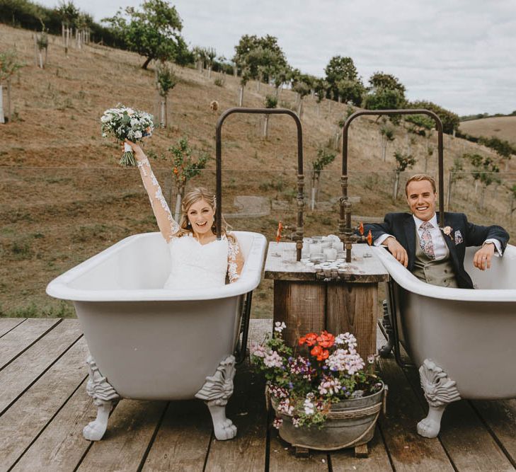 Bride in Lace Sincerity Bridal Wedding Dress and Groom in Blue Ted Baker Suit Sitting in Twin Rolltop Baths on The Old Piggyery Veranda at Devon Wedding Venue Windout Barn