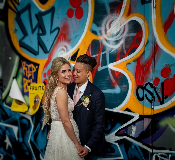 Bride in Navy Suit and Pink Tie and Bride in Flora Mila Wedding Dress Embracing in Front of a Wall Muriel