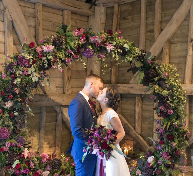 Bride in Jesus Peiro Wedding Dress and Groom in Blue  Hugo Boss Suit Standing in Front of a Floral Moon Gate