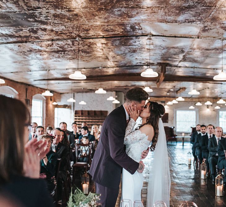 Bride in Lace Bardot Wedding Dress and Groom in a Moss Bros. Suit Kissing at the Altar