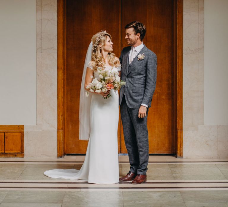 Bride in Lace Off The Shoulder Jennifer Regan Wedding Dress and Flower Crown and Groom in Grey Suit