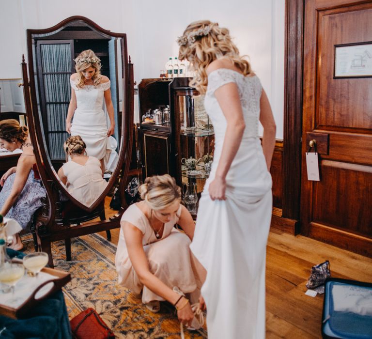 Bridesmaid in Peach Dress Helping Bride in Lace Off The Shoulder Jennifer Regan Wedding Dress Put On Her Harriet Wilde Wedding Bridal Shoes