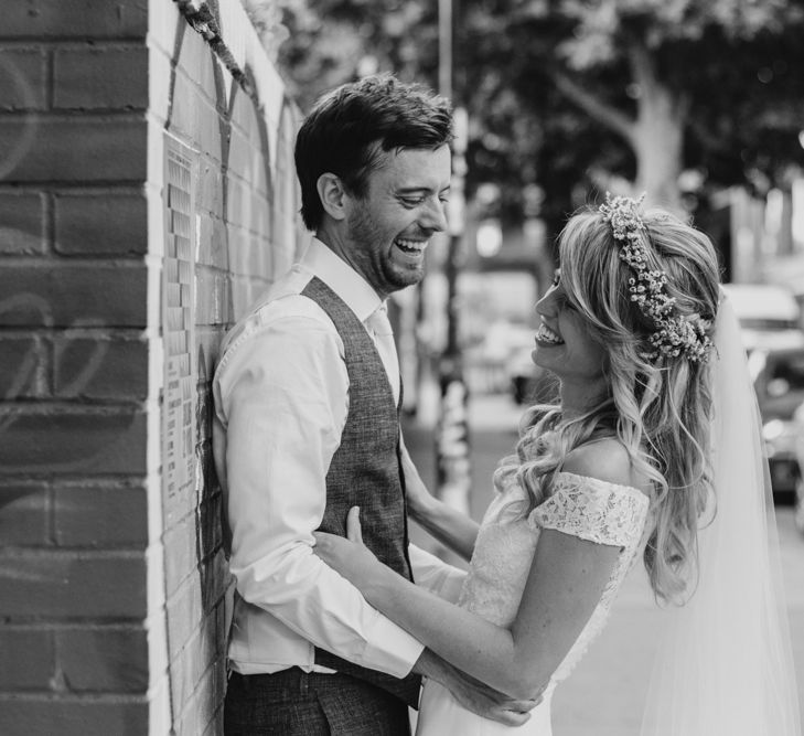 Bride in Lace Off The Shoulder Jennifer Regan Wedding Dress and Flower Crown and Groom in Grey Waistcoat Laughing