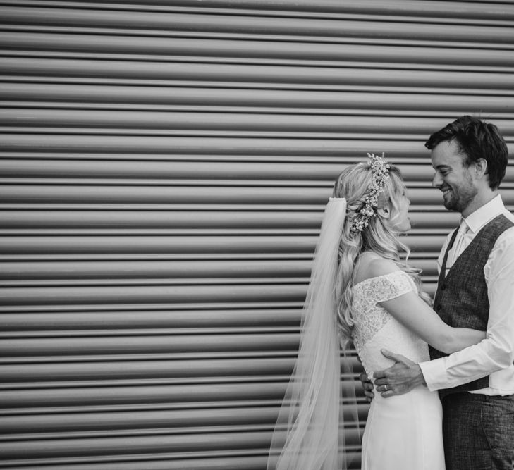 Bride in Lace Off The Shoulder Jennifer Regan Wedding Dress and Flower Crown and Groom in Grey Waistcoat Embracing