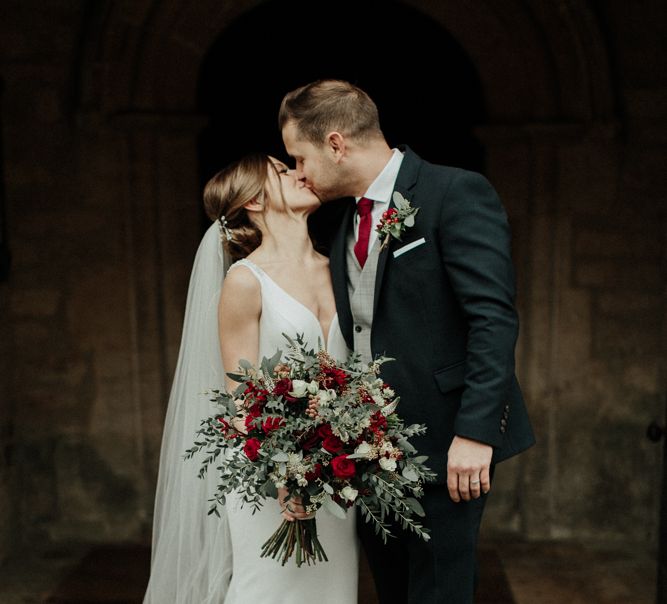 Bride in Essense of Australia Wedding Dress Holding a Red Flower and Foliage Bouquet and Groom in Marks and Spencer Suit Kissing Outside the Church