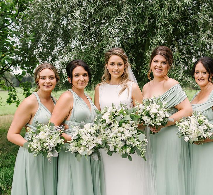 Bridal Party Portrait with Bridesmaids in Pale Green Multiway Dresses and Bride in Pronovias Wedding Dress Holding White Flower and Foliage Bouquets