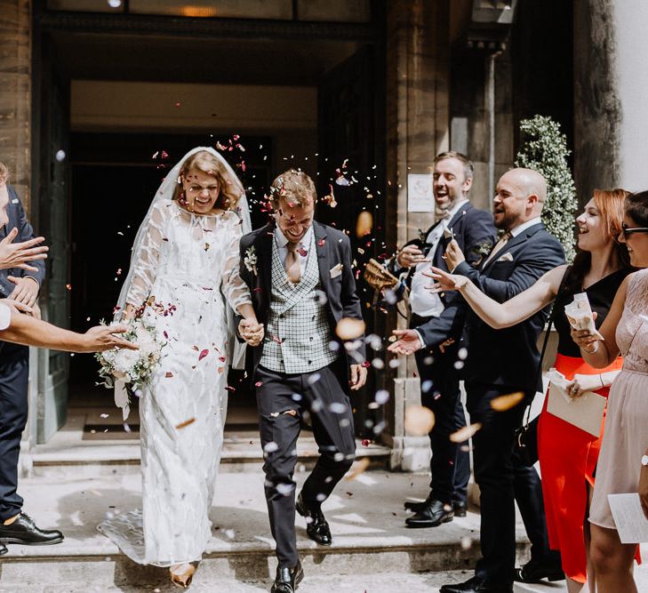 Bride in Embroidered Tulle Halfpenny London Dress with High Neckline, Batwing Sleeves and Low V-Back | Fingertip Length Veil from Florence &amp; Dot | Bridal Bouquet of White and Blush Pink Flowers with Eucalyptus and Trailing Silk Ribbon | Groom in Navy Slim Fit Suit with Checked Double Breasted Waistcoat and Blush Pink Knitted Tie | Confetti Moment at Stoke Newington Town Hall | Floral Arch and Bride in Batwing Sleeve Halfpenny Dress for London Pub Wedding | Caitlin + Jones