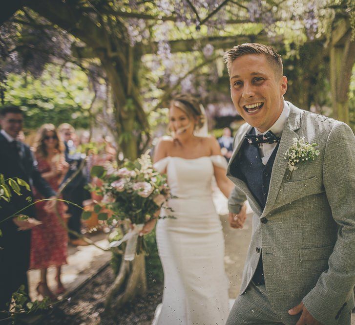 Confetti exit for bride in flat wedding shoes and groom in light suit with gypsophilia buttonhole