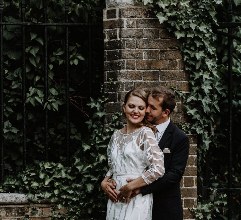 Bride in Embroidered Tulle Halfpenny London Dress with High Neckline, Batwing Sleeves and Low V-Back | Groom in Navy Slim Fit Suit with Checked Double Breasted Waistcoat and Blush Pink Knitted Tie | Floral Arch and Bride in Batwing Sleeve Halfpenny Dress for London Pub Wedding | Caitlin + Jones