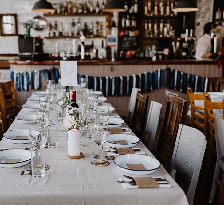 Banquet Tables | Mismatched Chairs | White Enamel Plates with Blue Rim | White Pillar Candle | Kraft Paper Wedding Breakfast Menus | Bud Vases with White Flowers and Eucalyptus | Blue and White Tassels | Floral Arch and Bride in Batwing Sleeve Halfpenny Dress for London Pub Wedding | Caitlin + Jones