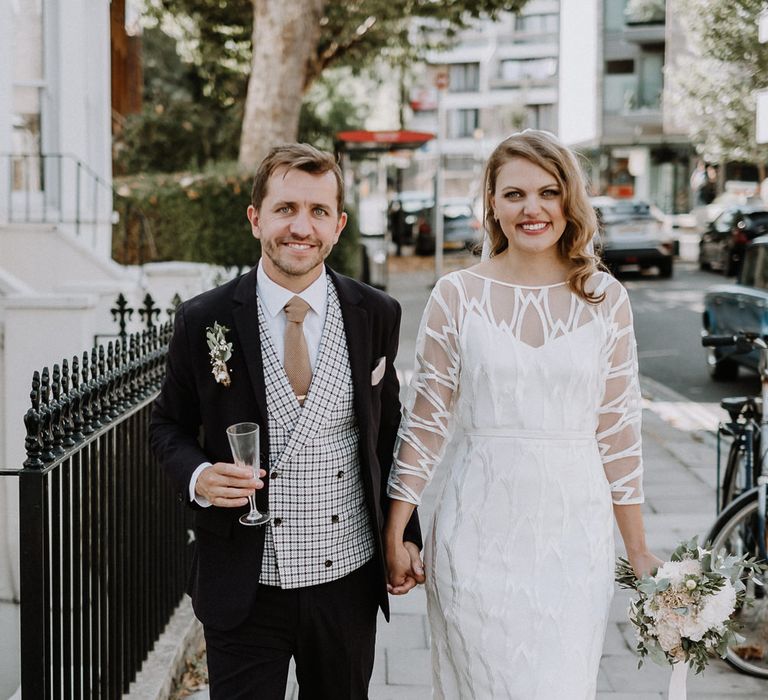 Bride in Embroidered Tulle Halfpenny London Dress with High Neckline, Batwing Sleeves and Low V-Back | Fingertip Length Veil from Florence &amp; Dot | Bridal Bouquet of White and Blush Pink Flowers with Eucalyptus and Trailing Silk Ribbon | Groom in Navy Slim Fit Suit with Checked Double Breasted Waistcoat and Blush Pink Knitted Tie | Floral Arch and Bride in Batwing Sleeve Halfpenny Dress for London Pub Wedding | Caitlin + Jones