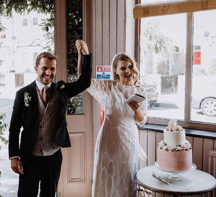 Bride in Embroidered Tulle Halfpenny London Dress with High Neckline, Batwing Sleeves and Low V-Back | Fingertip Length Veil from Florence &amp; Dot | Groom in Navy Slim Fit Suit with Checked Double Breasted Waistcoat and Blush Pink Knitted Tie | Two-Tier Wedding Cake with Buttercream Icing and Iced Flowers | Wedding Reception at The Star, Highgate | Floral Arch and Bride in Batwing Sleeve Halfpenny Dress for London Pub Wedding | Caitlin + Jones