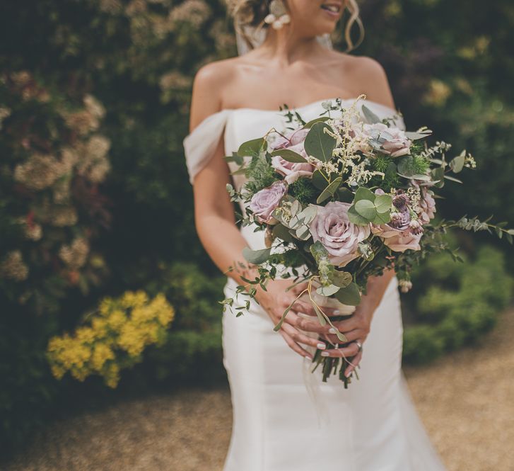 Bride in flat wedding shoes in off-the-shoulder wedding dress and beautiful lilac bouquet