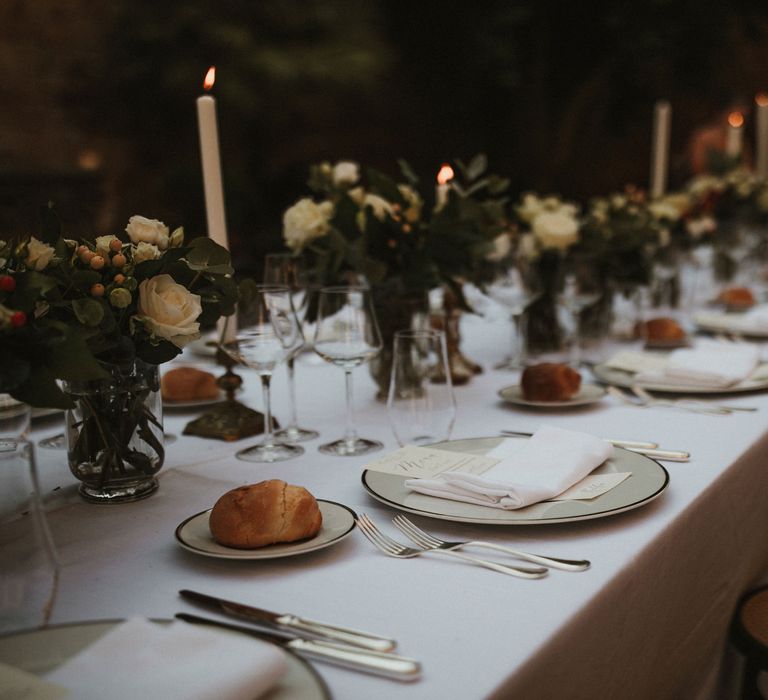 Wedding Breakfast Table in Courtyard at La Badia Di Orvieto Italy | White Tapered Candles | Gold Candlesticks | Hurricane Vases with White Flowers and Foliage | White Charger Plate with Gold Rim | Personalised Calligraphy Menu | Intimate Italian Castle Wedding with Prosecco Tower | James Frost Photography