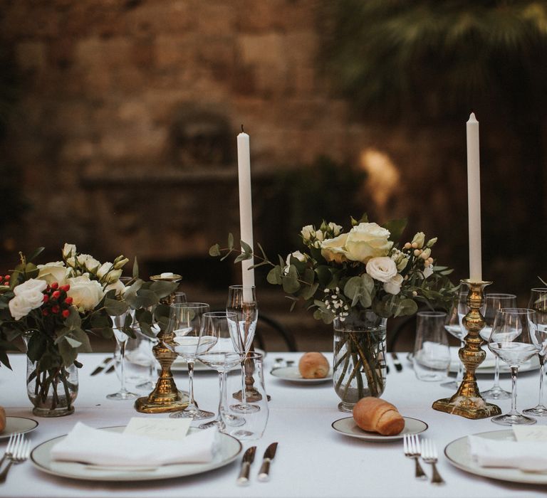 Wedding Breakfast Table in Courtyard at La Badia Di Orvieto Italy | White Tapered Candles | Gold Candlesticks | Hurricane Vases with White Flowers and Foliage | White Charger Plate with Gold Rim | Personalised Calligraphy Menu | Intimate Italian Castle Wedding with Prosecco Tower | James Frost Photography