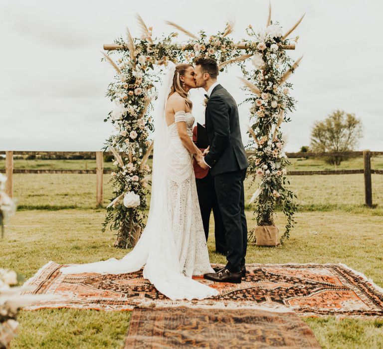 Boho Bride in Rue De Seine Wedding Dress and Groom in Black Tie Suit Kissing at the Floral Arch Altar