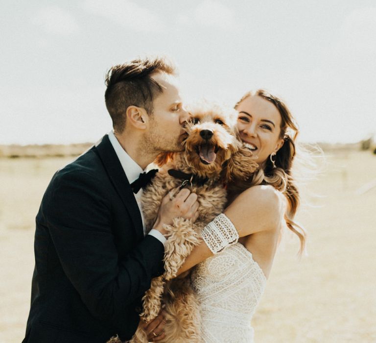 Boho Bride in Rue De Seine Wedding Dress and Groom in Black Tie Suit Hugging Their Pet Cockapoo