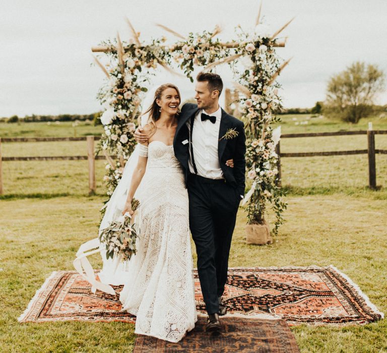 Boho Bride in Rue de Seine Wedding Dress and Groom in Black Tie Suit Embracing in Front of Their Floral Arch