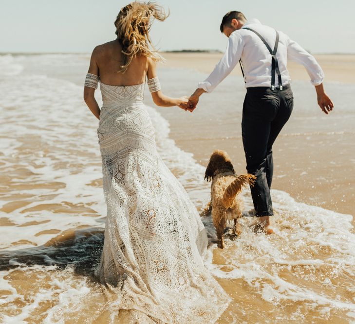 Boho Bride in Rue de Seine Wedding Dress and Groom in Black Tie Suit Walking Through The Surf with their Pet Cockapoo