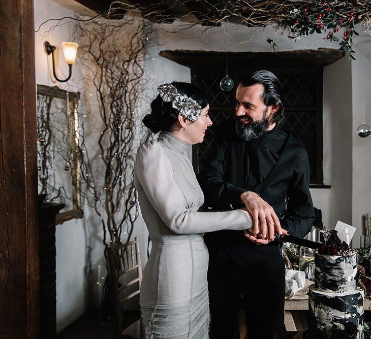 Bride and groom cut the cake at gothic wedding wearing silver dress, vintage floral headpiece and black suit