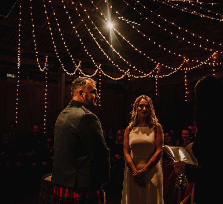 Bride in Anne Priscilla Signature Gown with Halterneck, Keyhole Back and Belt | Floor Length Veil | Groom in Mackintosh Tartan Red and Black Kilt with Green Jacket and Waistcoat | Kinkell Byre Wedding Venue | Festoon Light Canopy and Indoor Trees for Humanist Wedding in St Andrews | Carla Blain Photography