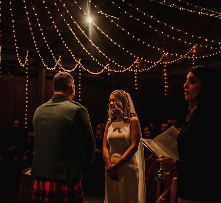 Bride in Anne Priscilla Signature Gown with Halterneck, Keyhole Back and Belt | Floor Length Veil | Groom in Mackintosh Tartan Red and Black Kilt with Green Jacket and Waistcoat | Kinkell Byre Wedding Venue | Festoon Light Canopy and Indoor Trees for Humanist Wedding in St Andrews | Carla Blain Photography