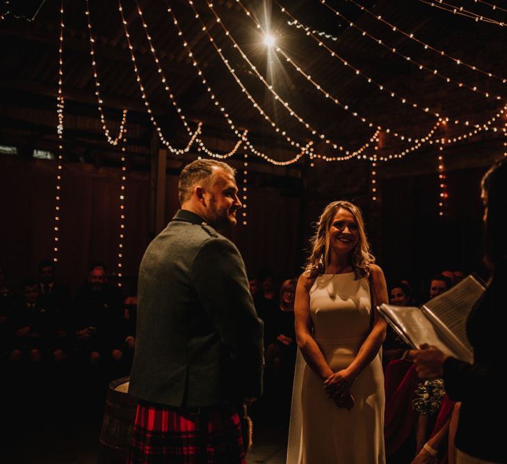 Bride in Anne Priscilla Signature Gown with Halterneck, Keyhole Back and Belt | Floor Length Veil | Groom in Mackintosh Tartan Red and Black Kilt with Green Jacket and Waistcoat | Kinkell Byre Wedding Venue | Festoon Light Canopy and Indoor Trees for Humanist Wedding in St Andrews | Carla Blain Photography