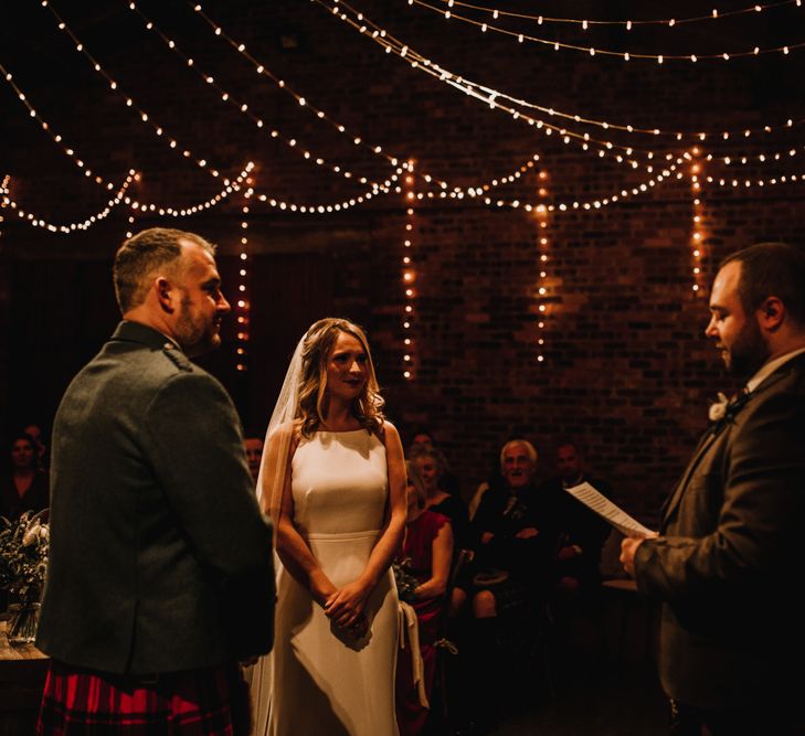 Bride in Anne Priscilla Signature Gown with Halterneck, Keyhole Back and Belt | Floor Length Veil | Groom in Mackintosh Tartan Red and Black Kilt with Green Jacket and Waistcoat | Kinkell Byre Wedding Venue | Festoon Light Canopy and Indoor Trees for Humanist Wedding in St Andrews | Carla Blain Photography