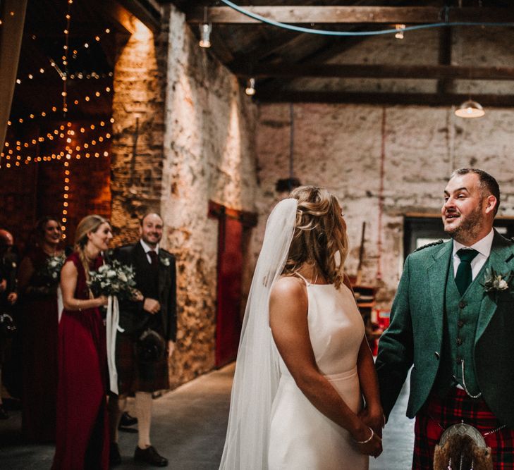 Bride in Anne Priscilla Signature Gown with Halterneck, Keyhole Back and Belt | Floor Length Veil | Groom in Mackintosh Tartan Red and Black Kilt with Green Jacket and Waistcoat | Kinkell Byre Wedding Venue | Festoon Light Canopy and Indoor Trees for Humanist Wedding in St Andrews | Carla Blain Photography