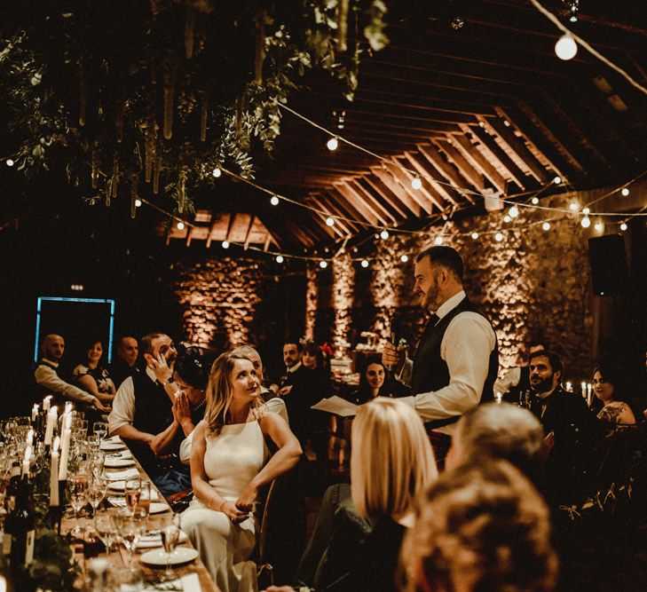 Bride in Anne Priscilla Signature Gown with Halterneck, Keyhole Back and Belt | Groom in Mackintosh Tartan Red and Black Kilt with Green Jacket and Waistcoat | Wooden Banquet Tables | Foliage Table Runners | White Tapered Candles | Foliage Hanging Decoration | Kinkell Byre Wedding Venue | Festoon Light Canopy and Indoor Trees for Humanist Wedding in St Andrews | Carla Blain Photography