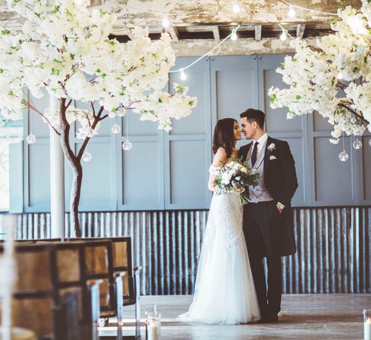 Bride in Fishtail Pronovias Wedding Dress with Off Shoulder Sleeves and Veil Cape | Groom in Navy Tails and Grey Waistcoat | Bride Wearing Hair Down | Bridal Bouquet of White Roses and Greenery | Cherry Blossom Tree Altar | Festoon Lights | Lace Bridal Cape Veil &amp; Fishtail Wedding Dress by Pronovias | On Love and Photography