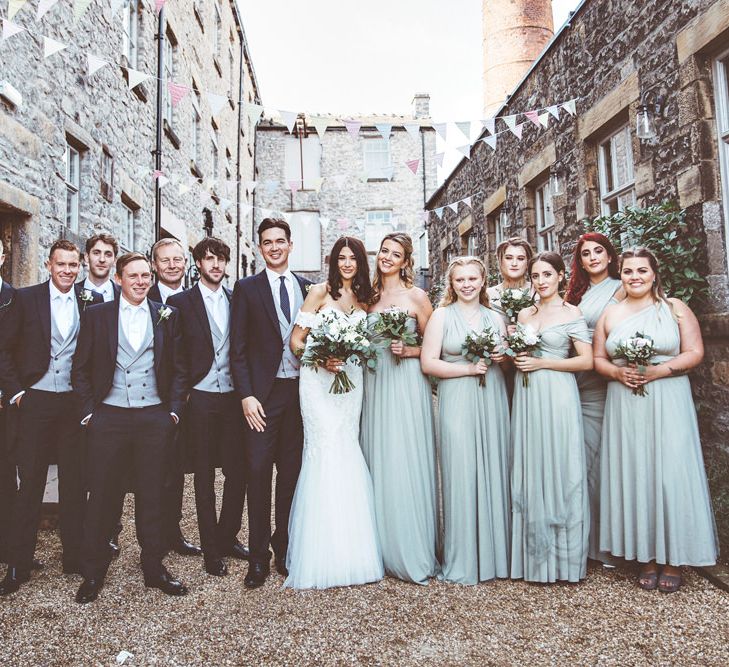 Bride in Fishtail Pronovias Wedding Dress with Off Shoulder Sleeves and Veil Cape | Bride Wearing Hair Down | Bridal Bouquet of White Roses and Greenery | Bridesmaids in Fossil Green Multiway Dresses | Bridesmaid Bouquets of Dusky Purple Roses and Greenery | Groom and Groomsmen in Navy Tails and Grey Waistcoats | Wedding Bunting | Lace Bridal Cape Veil &amp; Fishtail Wedding Dress by Pronovias | On Love and Photography