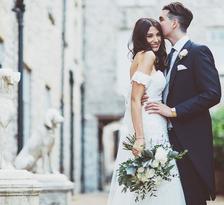 Bride in Fishtail Pronovias Wedding Dress with Off Shoulder Sleeves and Veil Cape | Groom in Navy Tails and Grey Waistcoat | Bride Wearing Hair Down | Bridal Bouquet of White Roses and Greenery | Lace Bridal Cape Veil &amp; Fishtail Wedding Dress by Pronovias | On Love and Photography