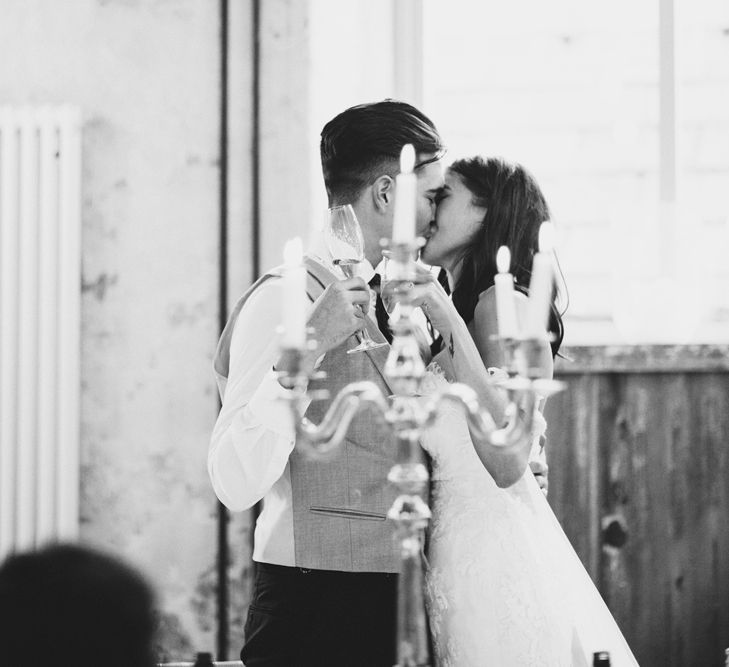 Newlywed Kiss | Bride in Fishtail Pronovias Wedding Dress with Off Shoulder Sleeves and Veil Cape | Groom in Navy Tails and Grey Waistcoat | Gold Candleabra with White Tapered Candles | Bride Wearing Hair Down | Exposed Copper Piping | Lace Bridal Cape Veil &amp; Fishtail Wedding Dress by Pronovias | On Love and Photography