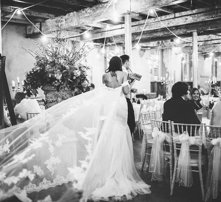 Bride in Fishtail Pronovias Wedding Dress with Off Shoulder Sleeves and Veil Cape | Groom in Navy Tails and Grey Waistcoat | Chalkboard Table Plan | White Material Chair Sashes | Oversized Floral Urn | Festoon Lights | Bride Wearing Hair Down | Lace Bridal Cape Veil &amp; Fishtail Wedding Dress by Pronovias | On Love and Photography