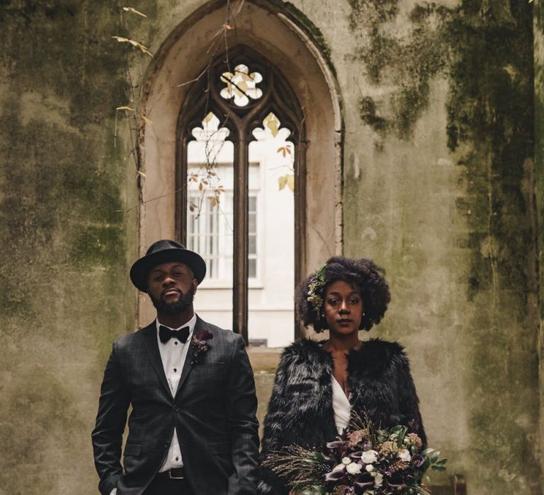 Bride in black faux fur cover up and groom in hat holding hands at St Dunstan in the East gardens