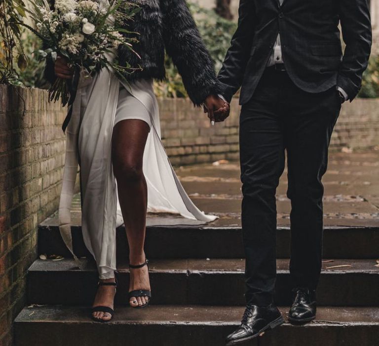 Bride in satin dress and faux fur coat holding hands with her groom