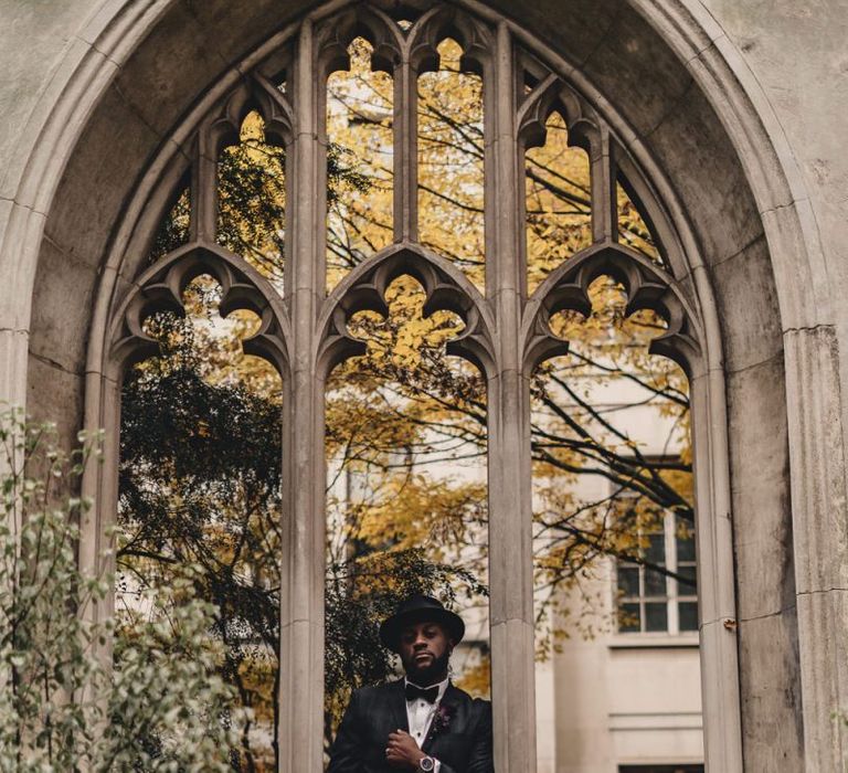 Stylish groom in bow tie and hat standing at St Dunstan in the East  church gardens