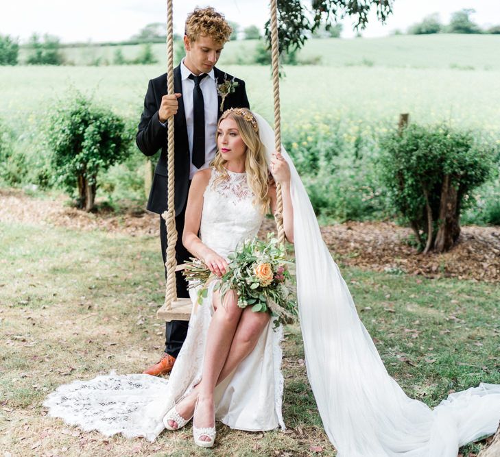 Bride in Lace Halterneck Wattens Wedding Dress with Front Slit and Groom in Black Suit and Tie Sitting on a Swing