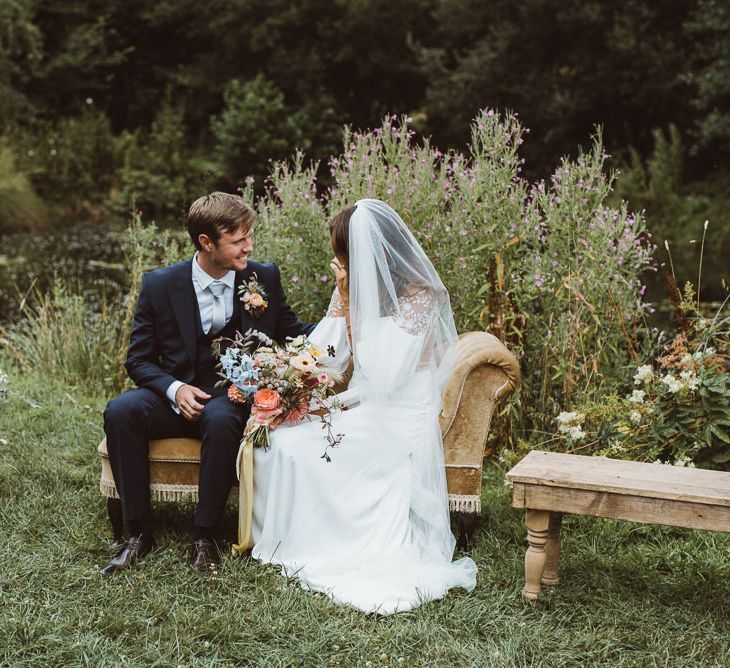 Bride in Rime Arodaky Wedding Dress and Groom is Reiss Suit Sitting on a Vintage Chaise Lounge During Outdoor Wedding Ceremony