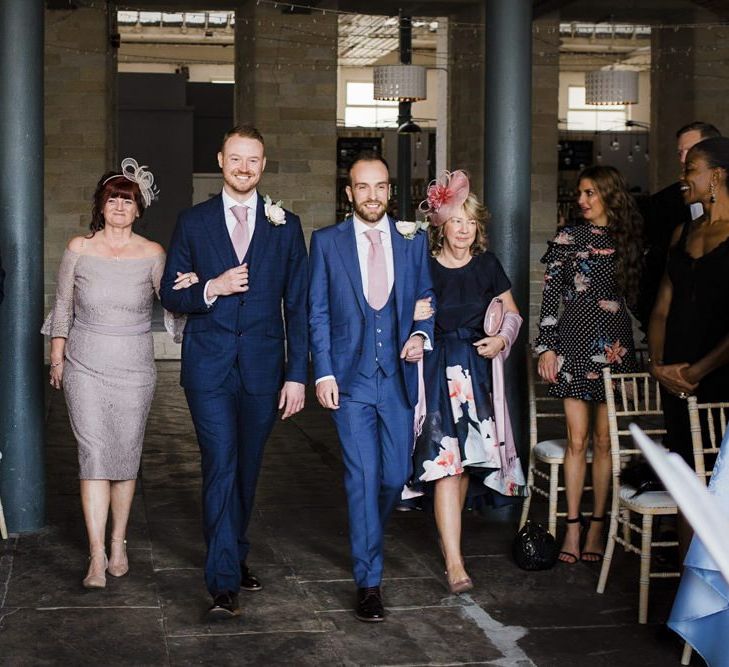 Two Grooms in Navy Blue Suits Walking Down the Aisle with Their Mums