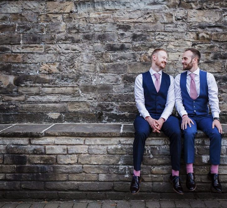 Two Grooms in Navy Suits with Pink Socks and Ties Sitting on a Wall