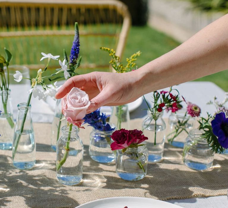 Person adding an antique pale pink rose to a top table flower arrangement made up of single stems in mini glass bottles