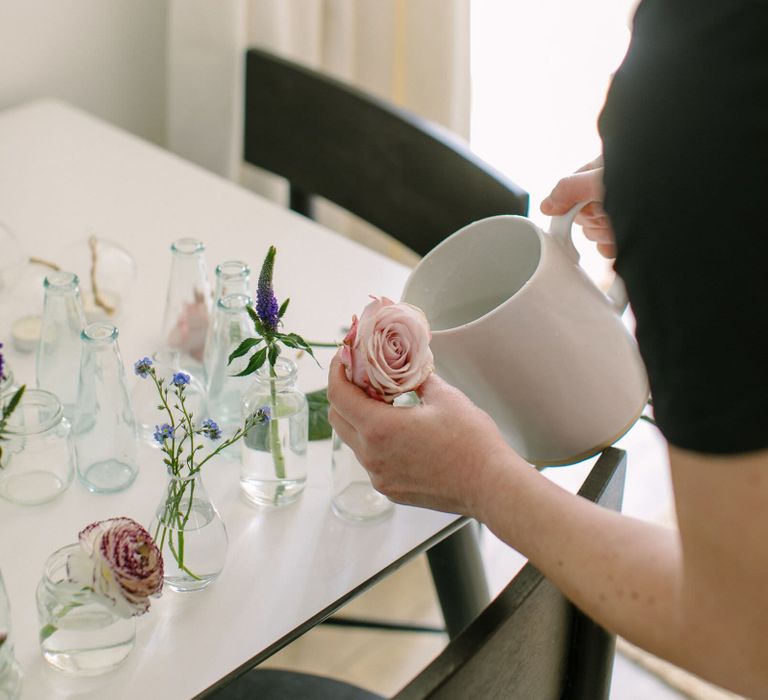 Person holding a pale pink antique rose and pouring water from a white ceramic jug into a small glass bottle vase