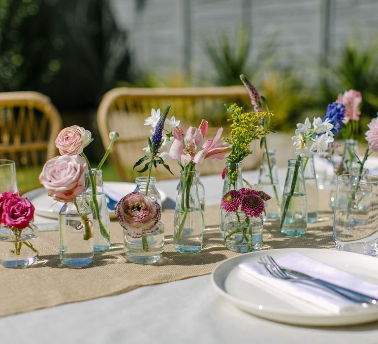 Table laid using neutral fabrics and tableware, and with a floral centrepiece made up of single stems in small glass vases