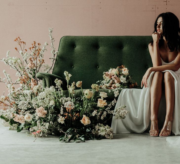 Bride in tulle wedding dress sitting on a forest green sofa with floral arrangement