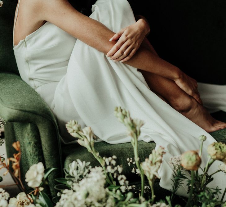 Bride curled up on a deep green velvet sofa decorated with muted flower arrangement