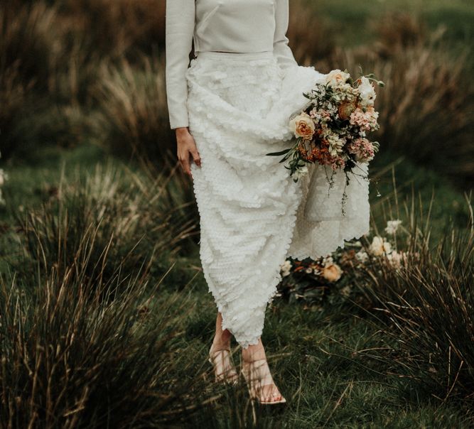 Bride in bridal crop top holding her bouquet and skirt in a field